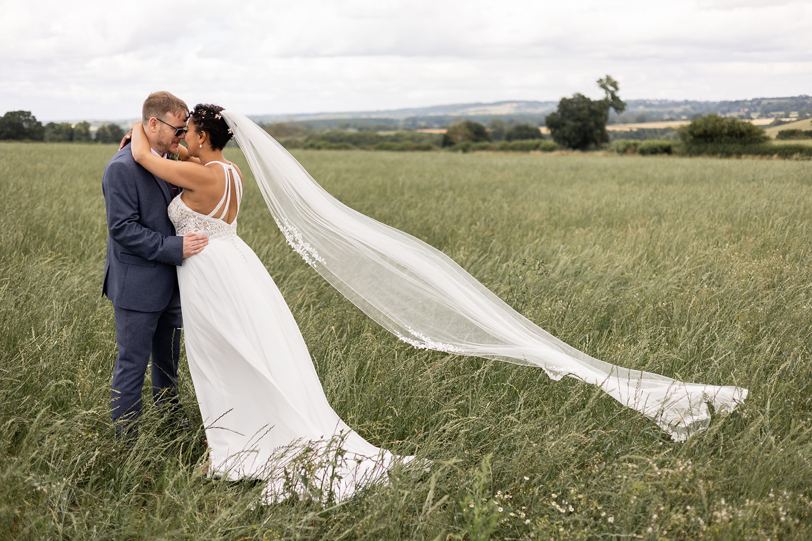 Couple in green field on wedding day with veil blowing in wind