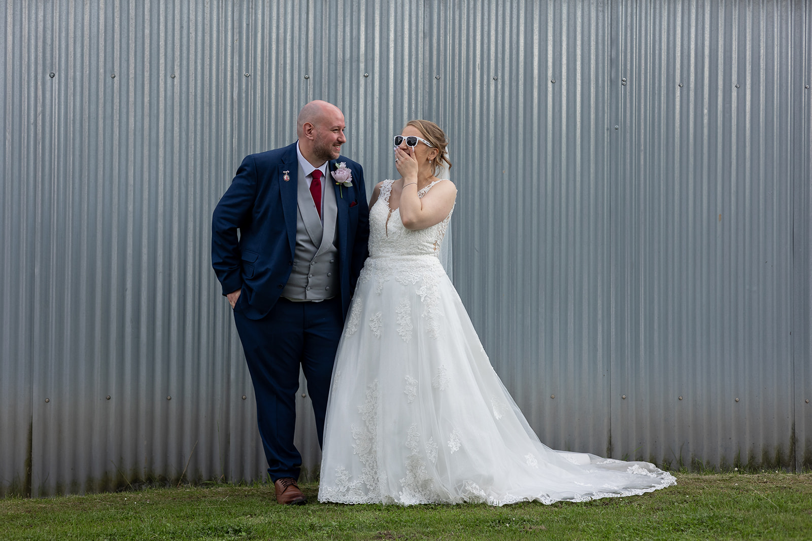 A wedding couple laughing in front of grey metal barn