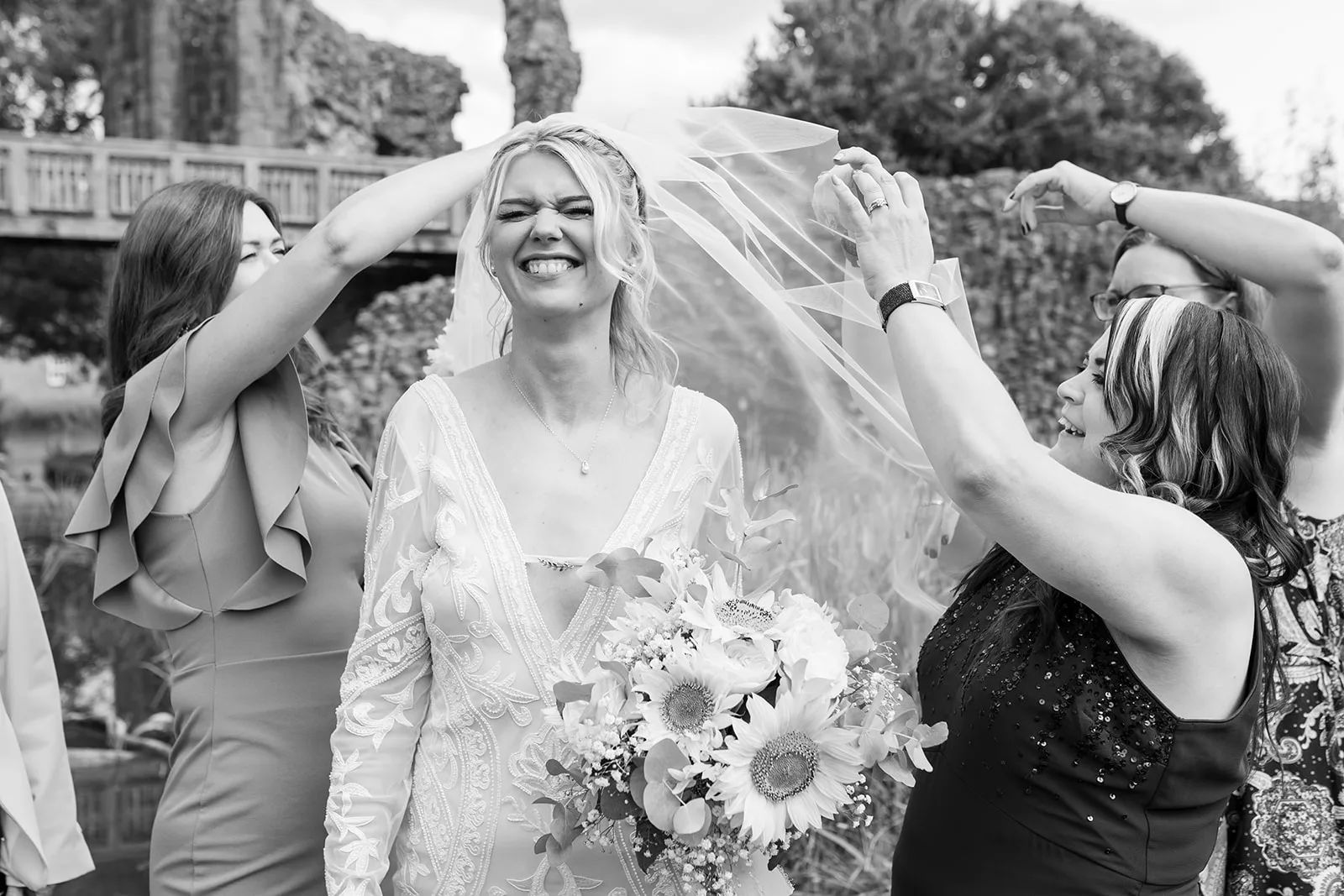 Black and white photo of a happy bride with a veil being lifted from her face. She is smiling.