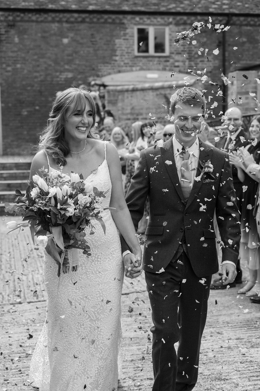 A black and white image of a couple on their wedding day smiling as they walk towards the camera through confetti