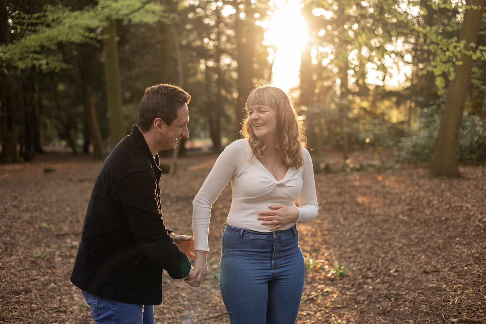 A couple are laughing in the woods together as the sun sets through the trees behind them. They look very happy.